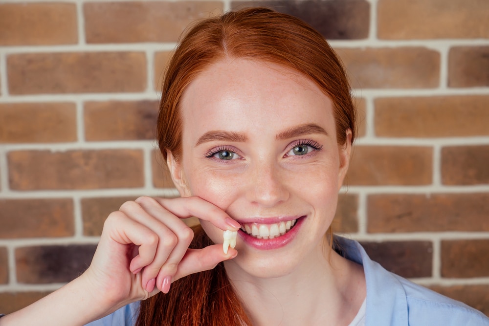 redhaired ginger female with snow-white smile holding white wisdom tooth after surgery removal of a tooth