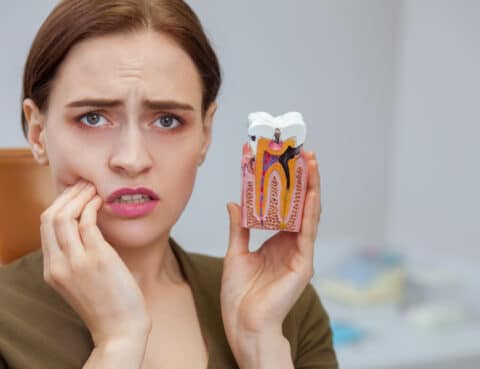 Young woman suffering from toothache, looking to the camera in despair, holding tooth mold showing cavity. Close up of a female patient with aching teeth visiting dental clinic. Dentistry, oral care