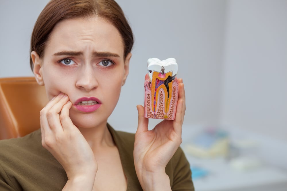 Young woman suffering from toothache, looking to the camera in despair, holding tooth mold showing cavity. Close up of a female patient with aching teeth visiting dental clinic. Dentistry, oral care