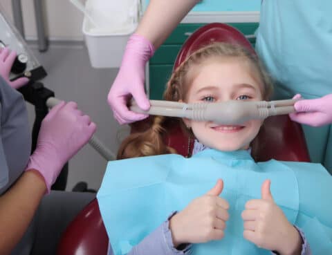 A little girl is comfortable to treat her teeth under superficial sedation. The girl smiles and holds two thumbs up. Milk teeth treatment.