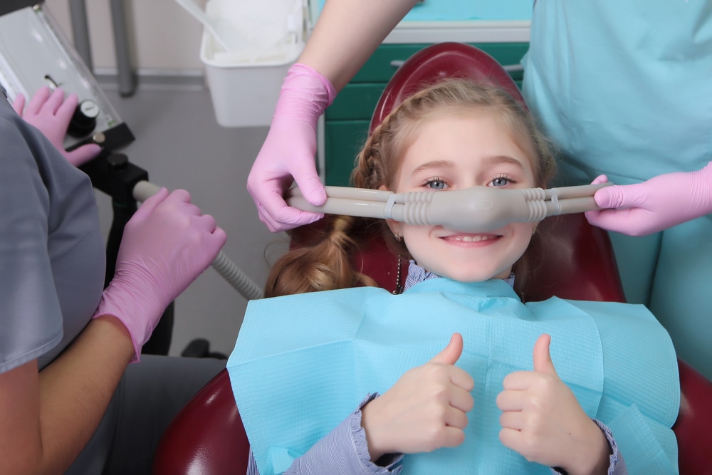 A little girl is comfortable to treat her teeth under superficial sedation. The girl smiles and holds two thumbs up. Milk teeth treatment.
