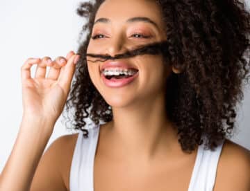 laughing african american girl with dental braces making fake mustache from hair, isolated on grey