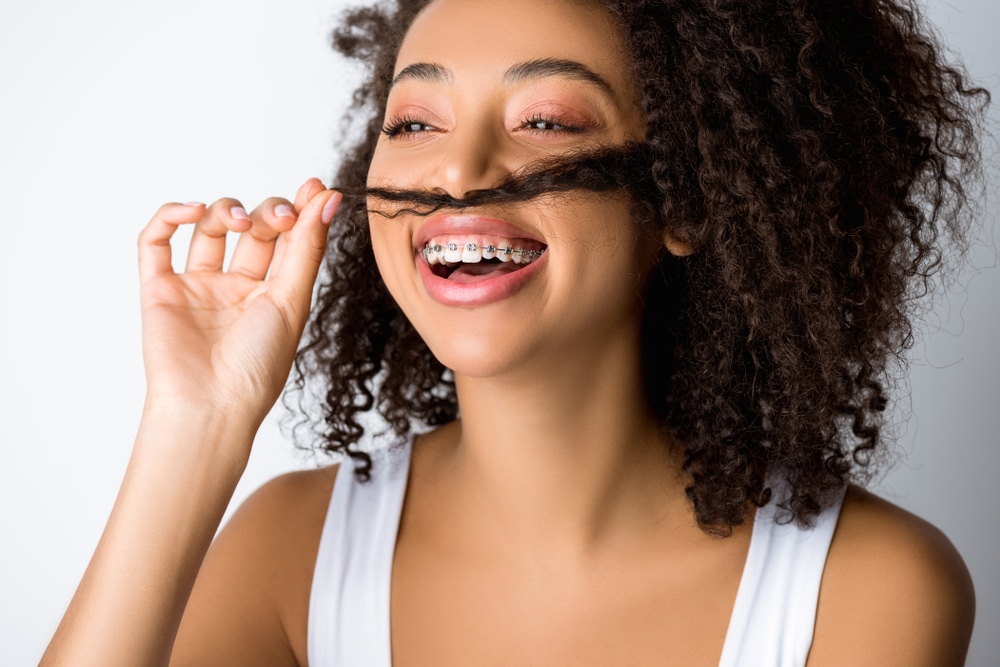 laughing african american girl with dental braces making fake mustache from hair, isolated on grey